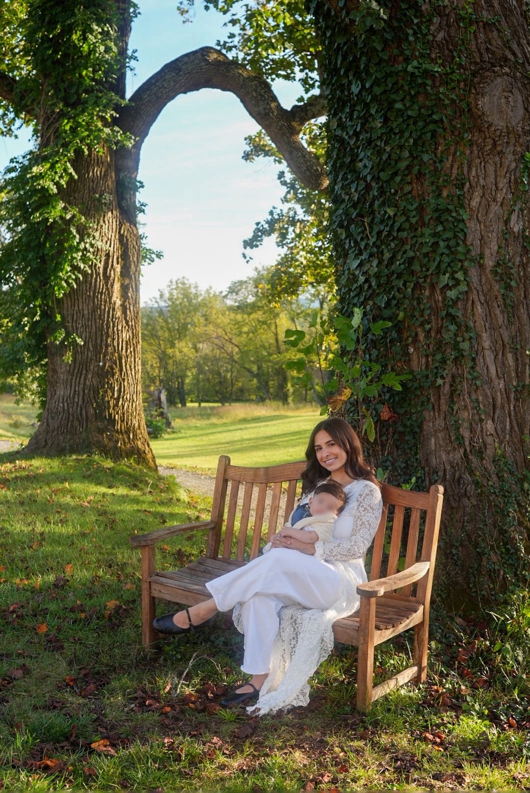 A mother holding her baby on a wooden bench beneath ivy-covered oak trees
