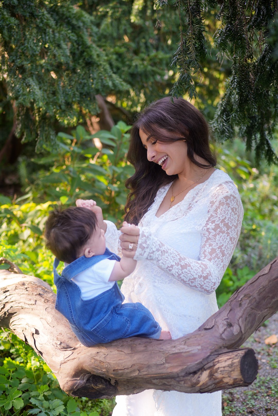 A mother laughing with her baby, who sits on a low tree branch