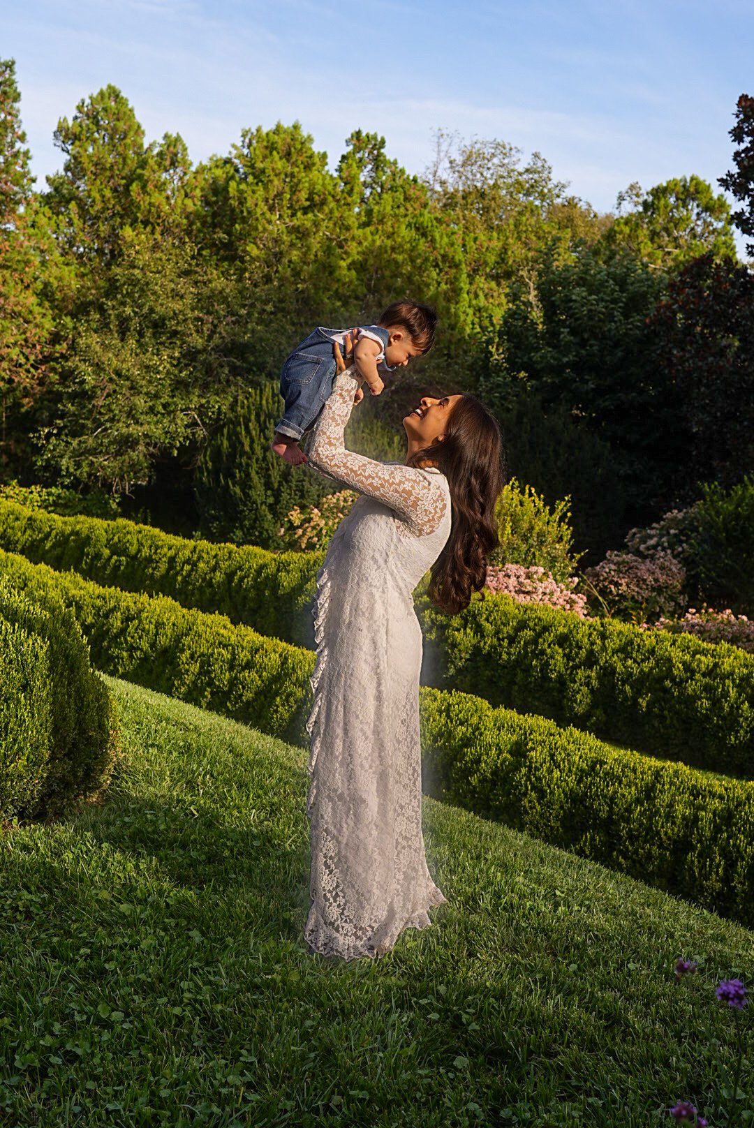 A mother in a white lace dress lifting her baby into the air in a hedged garden