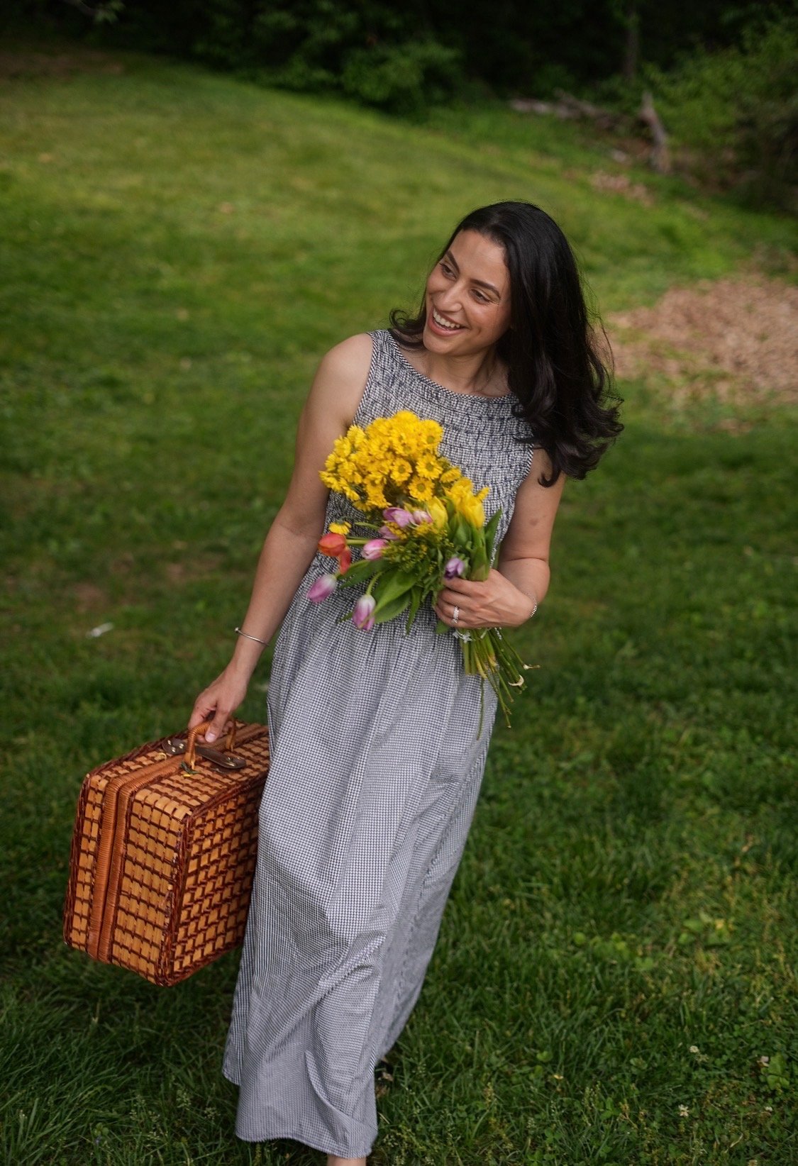 A woman laughing mid-stride in a meadow, carrying a picnic basket and a bouquet of wildflowers