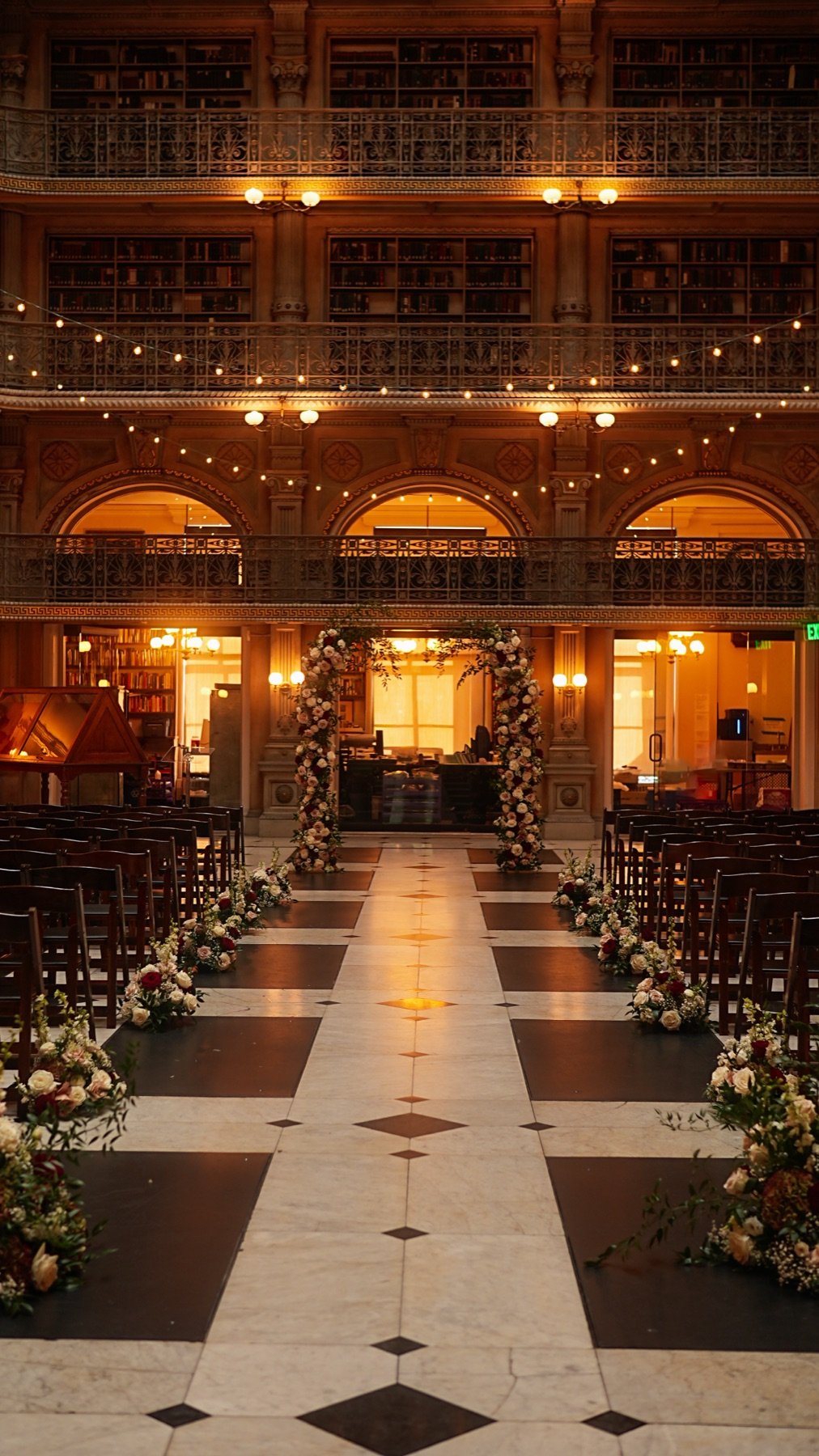 A ceremony aisle lined with flowers leading to a floral arch inside a grand library hall
