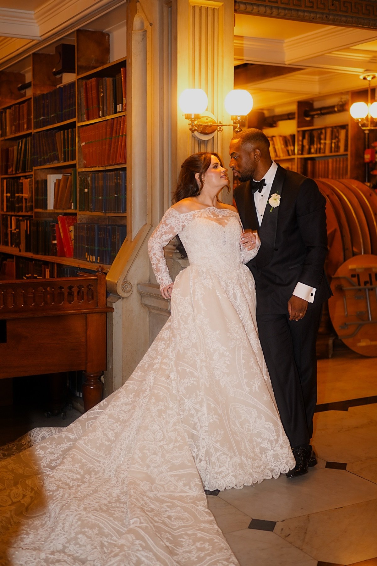 Bride and groom sharing a look beside tall library bookshelves, her lace train spread across the floor