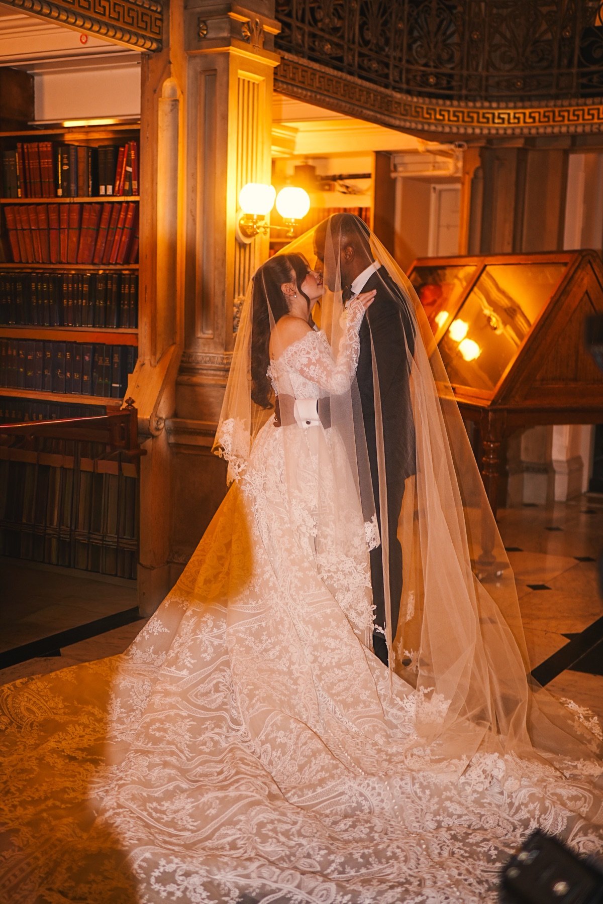 Bride and groom kissing beneath her veil beside the bookshelves of a historic library